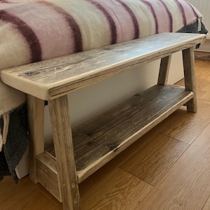 May include: A rectangular wooden bench featuring a weathered grey finish with visible wood grain patterns. The bench includes a flat top surface and a lower shelf supported by four angled legs, constructed with visible metal screws at the joints.