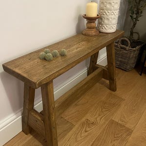 Op de afbeelding: Een rustieke houten bank met een natuurlijke bruine afwerking. De bank heeft een eenvoudig ontwerp met twee schuine poten aan elke kant. De bank staat op een hardhouten vloer.