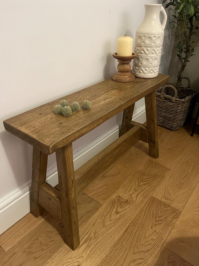 Op de afbeelding: Een rustieke houten bank met een natuurlijke bruine afwerking. De bank heeft een eenvoudig ontwerp met twee schuine poten aan elke kant. De bank staat op een hardhouten vloer.
