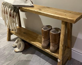 May include: A rustic wooden bench with a shelf underneath. The bench is made of reclaimed wood and has a natural finish. There are two pairs of brown boots and a pair of fuzzy slippers on the shelf. A white knit hat with a pom-pom sits on top of the bench.