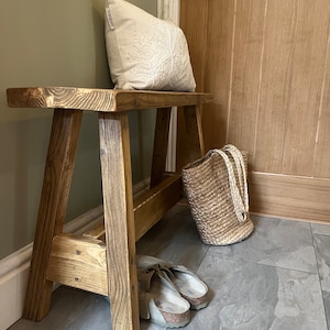 Op de afbeelding: Een rustieke houten bank met een natuurlijke afwerking. De bank heeft een eenvoudig ontwerp met twee poten aan elke kant en een brede zitting. Een wit kussen met een geborduurd ontwerp zit op de zitting. Een geweven mand en een paar sandalen staan op de vloer naast de bank.