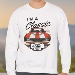May include: White sweatshirt with a classic car design in red, white, and brown. The text "I'M A Classic" is above the car, and the name "John" is below. The graphic is centrally placed on the chest.