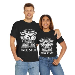 以下が含まれることがあります： 白いグラフィックデザインが施された黒いTシャツ2枚。グラフィックには、飛行機、ヤシの木、カクテル、そして「We're on our Honeymoon - Please give us free stuff」というテキストが描かれています。