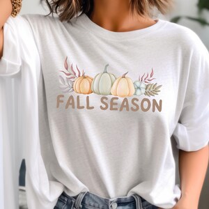 May include: White t-shirt with a graphic of three pumpkins, leaves, and flowers. The text "Fall Season" is printed below the pumpkins.