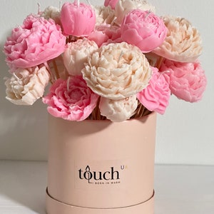 Pode incluir: Um buqu&ecirc; de velas em forma de pe&ocirc;nia em tons de rosa e creme, dispostas em uma caixa redonda rosa claro. A caixa tem um r&oacute;tulo que diz "touch" e "BORN IN WARM". As velas est&atilde;o em palitos de madeira.