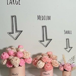 Pode incluir: Tr&ecirc;s arranjos florais em caixas redondas rosa com as etiquetas "Large", "Medium" e "Small". Os arranjos apresentam flores rosa e creme. As caixas t&ecirc;m uma faixa dourada e a palavra "touch" impressa.