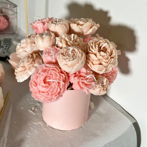 Pode incluir: Um buqu&ecirc; de velas em forma de pe&ocirc;nia em tons de rosa e p&ecirc;ssego. As velas s&atilde;o dispostas em um recipiente cil&iacute;ndrico rosa claro. Cada vela tem um pavio. O arranjo est&aacute; em uma superf&iacute;cie clara.