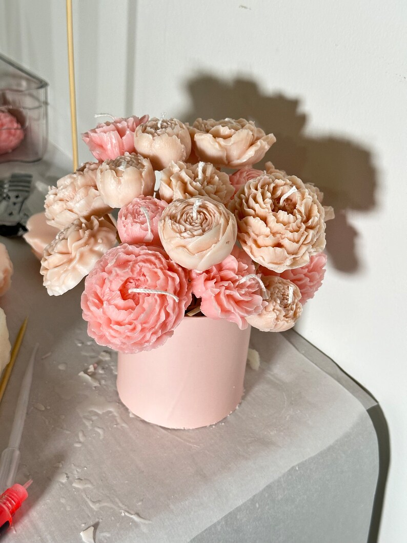Pode incluir: Um buqu&ecirc; de velas em forma de pe&ocirc;nia em tons de rosa e p&ecirc;ssego. As velas s&atilde;o dispostas em um recipiente cil&iacute;ndrico rosa claro. Cada vela tem um pavio. O arranjo est&aacute; em uma superf&iacute;cie clara.
