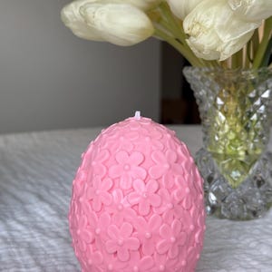 Puede incluir: Una vela rosa en forma de huevo con un diseño floral. La vela está cubierta de pequeñas flores rosas.