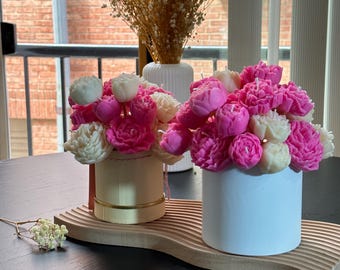 Peut inclure: Deux contenants en céramique blanche remplis de fleurs de cire roses et blanches. Les contenants sont posés sur un plateau en bois avec un motif ondulé.