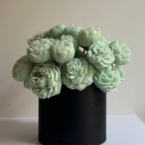 Pode incluir: Um buqu&ecirc; de velas em forma de pe&ocirc;nias verde s&aacute;lvia com pavios brancos, dispostas em um recipiente cil&iacute;ndrico preto. As velas s&atilde;o detalhadas para se assemelhar &agrave;s p&eacute;talas da flor. O arranjo est&aacute; sobre uma superf&iacute;cie branca.