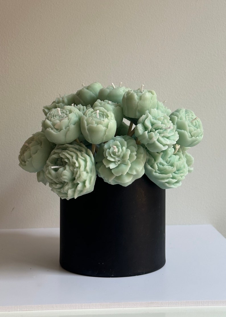 Pode incluir: Um buqu&ecirc; de velas em forma de pe&ocirc;nias verde s&aacute;lvia com pavios brancos, dispostas em um recipiente cil&iacute;ndrico preto. As velas s&atilde;o detalhadas para se assemelhar &agrave;s p&eacute;talas da flor. O arranjo est&aacute; sobre uma superf&iacute;cie branca.