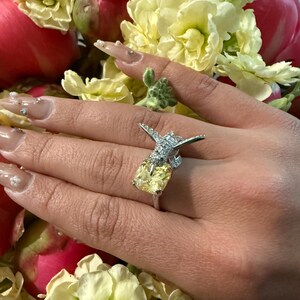 May include: A silver ring with a large, square, yellow gemstone and a decorative, sparkling accent. The ring is set against a backdrop of yellow and pink flowers.