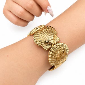 Könnte beinhalten: Ein goldfarbenes Armband mit einer Reihe von miteinander verbundenen, detailreichen Jakobsmuschel-Motiven. Die metallischen Glieder bilden ein durchgehendes Band und weisen eine polierte, reflektierende Oberfläche mit tiefen Rillen auf.