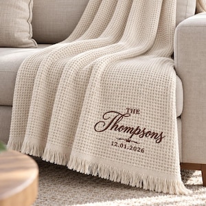 May include: A cream-coloured waffle-weave throw blanket featuring a fringed edge and brown embroidered text that reads THE Thompsons followed by the date 12.01.2026 and a small crown icon centred above the family name on the lower corner.