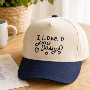Op de afbeelding: Een crèmekleurige baseballpet met een donkerblauwe klep en een donkerblauw geborduurd ontwerp op de voorkant met de tekst I L.O.V.E. Y.O.U. Daddy, omringd door verschillende kleine geborduurde hartjes in dezelfde donkerblauwe draad.