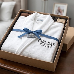 Könnte beinhalten: Ein weißes Spa-Geschenkset in einer braunen Box. Das Set enthält einen weißen Bademantel mit der Aufschrift "GIRL DAD EST. 2026", einen Kamm und ein Stück Seife. Ein blaues Band und ein Etikett mit der Aufschrift "DADA'S SPA DAY" sind um den Bademantel gebunden.