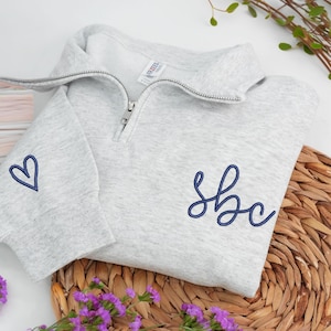 Könnte beinhalten: Hellgraues Sweatshirt mit kurzem Reißverschluss und marineblauen, gestickten Initialen und einem Herz. Das Sweatshirt liegt gefaltet auf einem geflochtenen Korb, mit lila Blumen im Vordergrund.