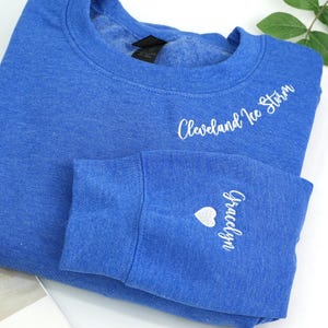 May include: A blue sweatshirt with the words "Cleveland Ice Storm" embroidered in white script across the chest. The sleeve cuff features the name "Gracelyn" and a heart symbol, also in white.