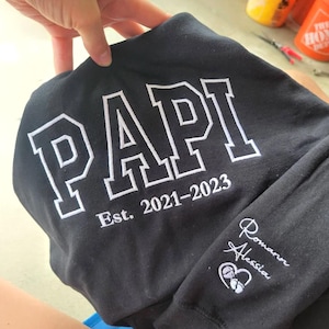 Puede incluir: Sudadera negra con la palabra "PAPI" en letras blancas grandes. Debajo, el texto "Est. 2021-2023". La manga tiene los nombres "Romann Alessia" bordados en blanco con un corazón y huellas de pies de bebé.