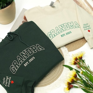May include: Two sweatshirts, one dark green and one cream-colored, with the words "GRANDPA" and "GRANDMA" embroidered in white and green, respectively. Both sweatshirts have "EST. 2025" and a name with a heart on the sleeve.