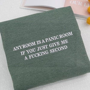 May include: A folded, sage green sweatshirt with white embroidered text. The text reads: "ANYROOM IS A PANIC ROOM IF YOU JUST GIVE ME A FUCKING SECOND". The sweatshirt is made of a soft material.