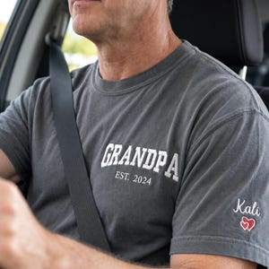 May include: A charcoal gray short-sleeved t-shirt featuring white embroidered text on the chest that reads "GRANDPA EST. 2024". The left sleeve displays the embroidered name "Kali" in white script above a small red heart outline design.