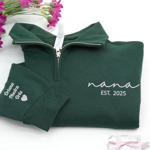 May include: A dark green quarter-zip sweatshirt featuring the word "nana" and "EST. 2025" in white script. The left cuff has embroidered names and a heart. The sweatshirt is folded, with a zipper and a tag.