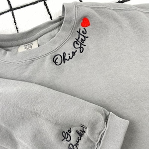 May include: A gray sweatshirt with the words "Ohio State" embroidered in black script, a red heart, and "Go Bucks!!" on the sleeve. The sweatshirt is made of a soft material and has a crew neck.
