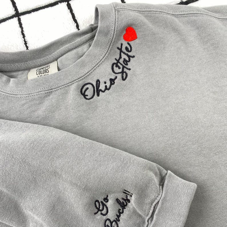 May include: A gray sweatshirt with the words "Ohio State" embroidered in black script, a red heart, and "Go Bucks!!" on the sleeve. The sweatshirt is made of a soft material and has a crew neck.