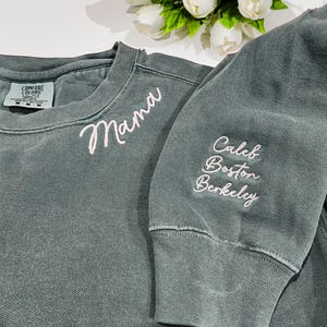 Blue spruce Comfort Colors sweatshirt with “Mama” embroidered in white script on the chest and personalized sleeve featuring the names “Caleb, Boston, Berkeley” in matching script. Cozy and stylish custom crewneck perfect for Mother’s Day, birthdays, or Christmas gifts. High-quality embroidery makes this sweatshirt a cherished keepsake, ideal for Etsy shoppers seeking personalized family apparel.