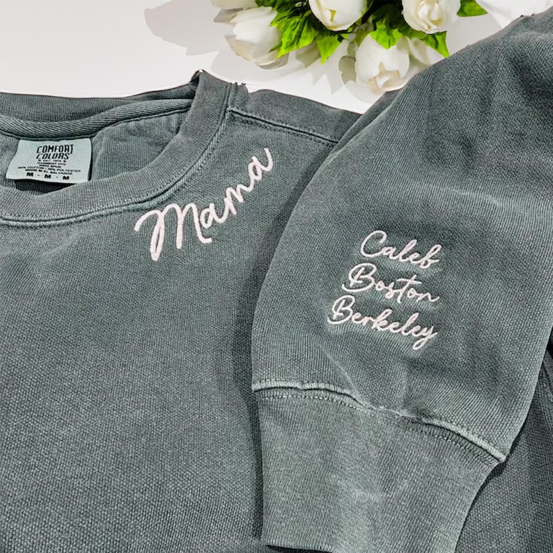 Blue spruce Comfort Colors sweatshirt with “Mama” embroidered in white script on the chest and personalized sleeve featuring the names “Caleb, Boston, Berkeley” in matching script. Cozy and stylish custom crewneck perfect for Mother’s Day, birthdays, or Christmas gifts. High-quality embroidery makes this sweatshirt a cherished keepsake, ideal for Etsy shoppers seeking personalized family apparel.