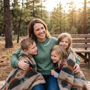 May include: A woman and three children are seated on a wooden bench outdoors. The woman wears a green sweatshirt with "mama" embroidered on it. The children are wrapped in patterned blankets, smiling. The background features a forest.
