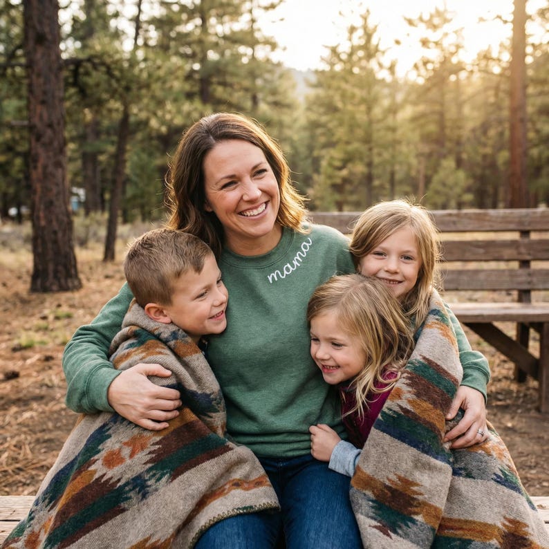 May include: A woman and three children are seated on a wooden bench outdoors. The woman wears a green sweatshirt with "mama" embroidered on it. The children are wrapped in patterned blankets, smiling. The background features a forest.