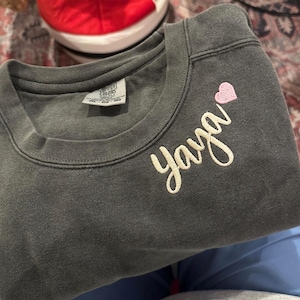 Pepper grey Comfort Colors sweatshirt with “Yaya” embroidered in cream script and a pink heart on the chest. Cozy and meaningful gift for grandmothers, Mother’s Day, birthdays, or Christmas. High-quality custom embroidery adds a personal touch, making this crewneck a cherished keepsake. Perfect personalized sweatshirt for Etsy shoppers seeking unique family gifts.