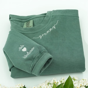 May include: A sage green sweatshirt with white embroidered text that reads "grammy" across the chest. The left sleeve has the names "Arlo Windsarose" with a heart symbol. The sweatshirt is folded on a white surface.