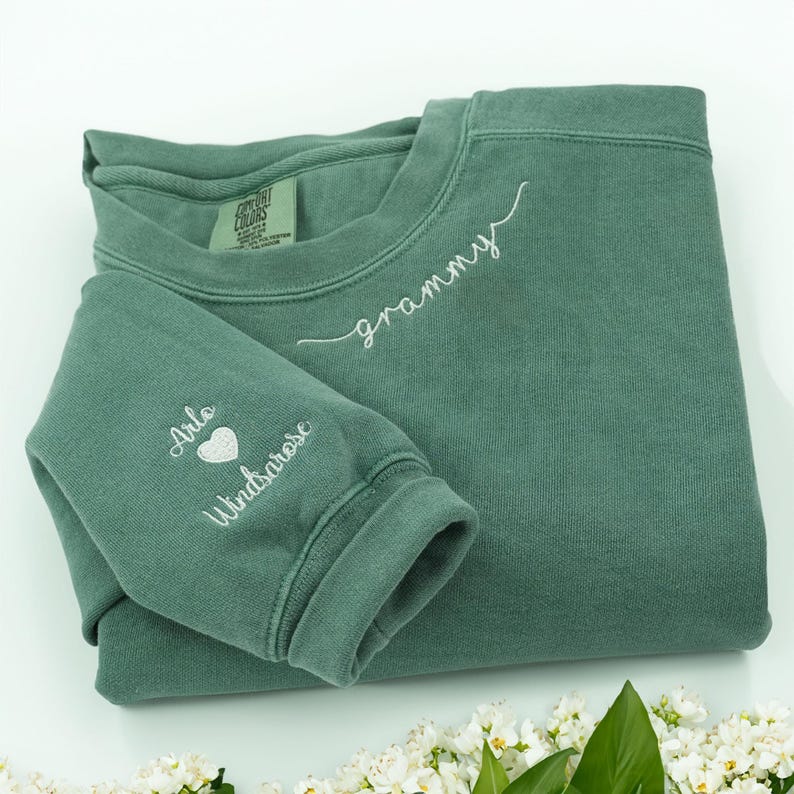 May include: A sage green sweatshirt with white embroidered text that reads "grammy" across the chest. The left sleeve has the names "Arlo Windsarose" with a heart symbol. The sweatshirt is folded on a white surface.
