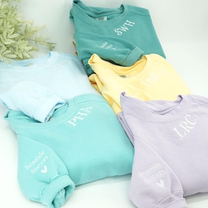 May include: A collection of pastel-colored sweatshirts, including light blue, teal, yellow, and lavender. Each sweatshirt features embroidered initials on the front and the words "Seaside Sisters" on the sleeve. The sleeves are folded, and the sweatshirts are stacked.