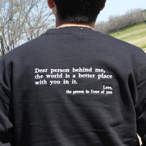 Op de afbeelding: Zwart sweatshirt met witte tekst op de rug. De tekst luidt: "Lieve persoon achter me, de wereld is een betere plek met jou erin. Liefs, de persoon voor je."