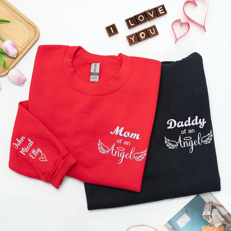 Red and black embroidered sweatshirts with “Mom of an Angel” and “Daddy of an Angel” text featuring angel wings and halo design. The red shirt sleeve is personalized with names and a heart, styled with tulips and chocolate letters spelling “I LOVE YOU.” Perfect memorial gift for parents honoring their angel baby.