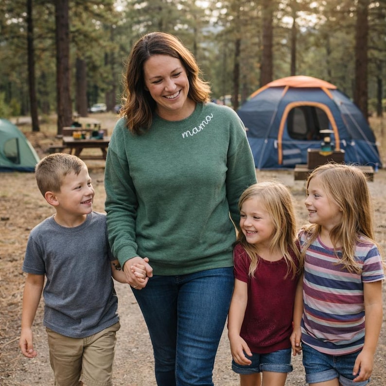May include: A woman wearing a green sweatshirt with the word "mama" embroidered on it, walking with three children. The children are wearing casual clothes. A blue tent is visible in the background, suggesting a camping trip.