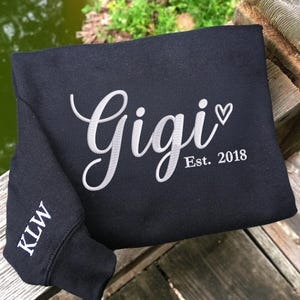 May include: Black sweatshirt with the name "Gigi" embroidered in white script, a small heart, and "Est. 2018." The left sleeve cuff displays the initials "KLW" in white. The sweatshirt is folded on a wooden surface.