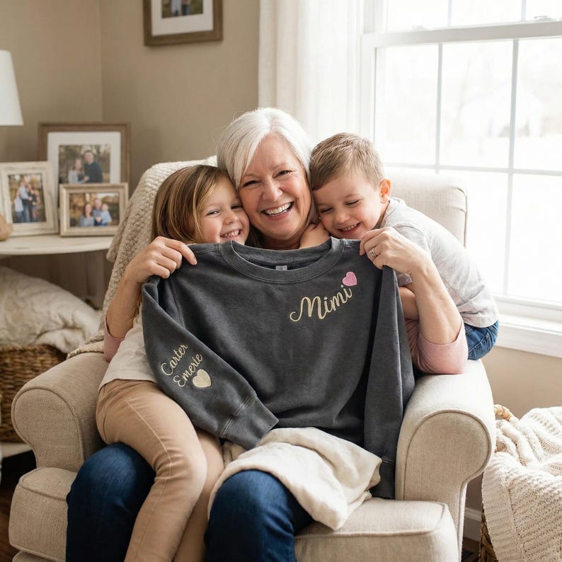 May include: A dark gray sweatshirt with the word "Mimi" in cursive script and a pink heart. The left sleeve has the names "Carter" and "Emerie" with a heart. The sweatshirt is being held by a woman and two children.