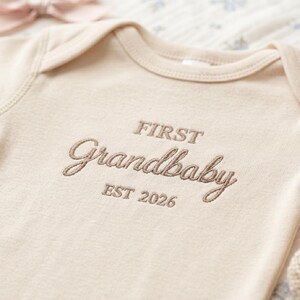 Detailed shot of the professional embroidery on a Natural-colored Rabbit Skins 4400 baby bodysuit. The text "First Grandbaby EST 2026" is stitched in a sophisticated script font. Highlighting the durable ribbed texture and precise needlework, this personalized baby clothing is an ideal keepsake for baby showers and gender-neutral nurseries.