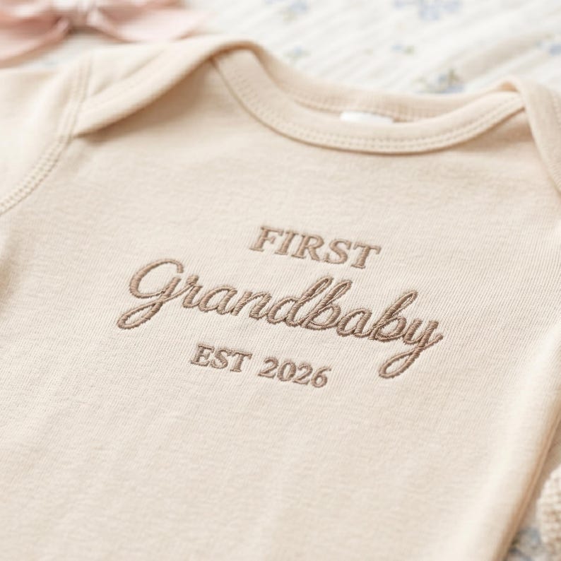 Detailed shot of the professional embroidery on a Natural-colored Rabbit Skins 4400 baby bodysuit. The text "First Grandbaby EST 2026" is stitched in a sophisticated script font. Highlighting the durable ribbed texture and precise needlework, this personalized baby clothing is an ideal keepsake for baby showers and gender-neutral nurseries.