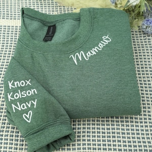 May include: A green sweatshirt with white text that reads "Mama" on the chest and "Knox, Kolson, Navy" on the sleeve, with a heart symbol. The sweatshirt is folded on a patterned surface.