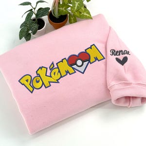 May include: A light pink sweatshirt with the embroidered word "Pokémon" in yellow and blue, with a heart-shaped Poké Ball design. The sleeve has the name "Renae" and a black heart embroidered on it.