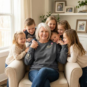 May include: A smiling older woman wearing a gray sweatshirt with the word "Mimi" in cursive script and a pink heart. She is surrounded by six children. The sweatshirt also has the names "Carter" and "Emery" on the sleeve.