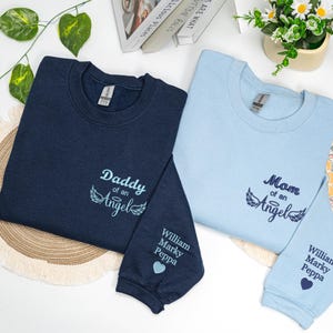 Matching navy and light blue embroidered sweatshirts with “Mom of an Angel” and “Daddy of an Angel” designs, each featuring angel wings and personalized names on the sleeve beside a small heart. A custom memorial gift honoring an angel baby, ideal for comfort and remembrance.