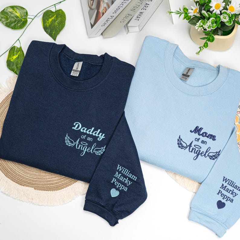 Matching navy and light blue embroidered sweatshirts with “Mom of an Angel” and “Daddy of an Angel” designs, each featuring angel wings and personalized names on the sleeve beside a small heart. A custom memorial gift honoring an angel baby, ideal for comfort and remembrance.